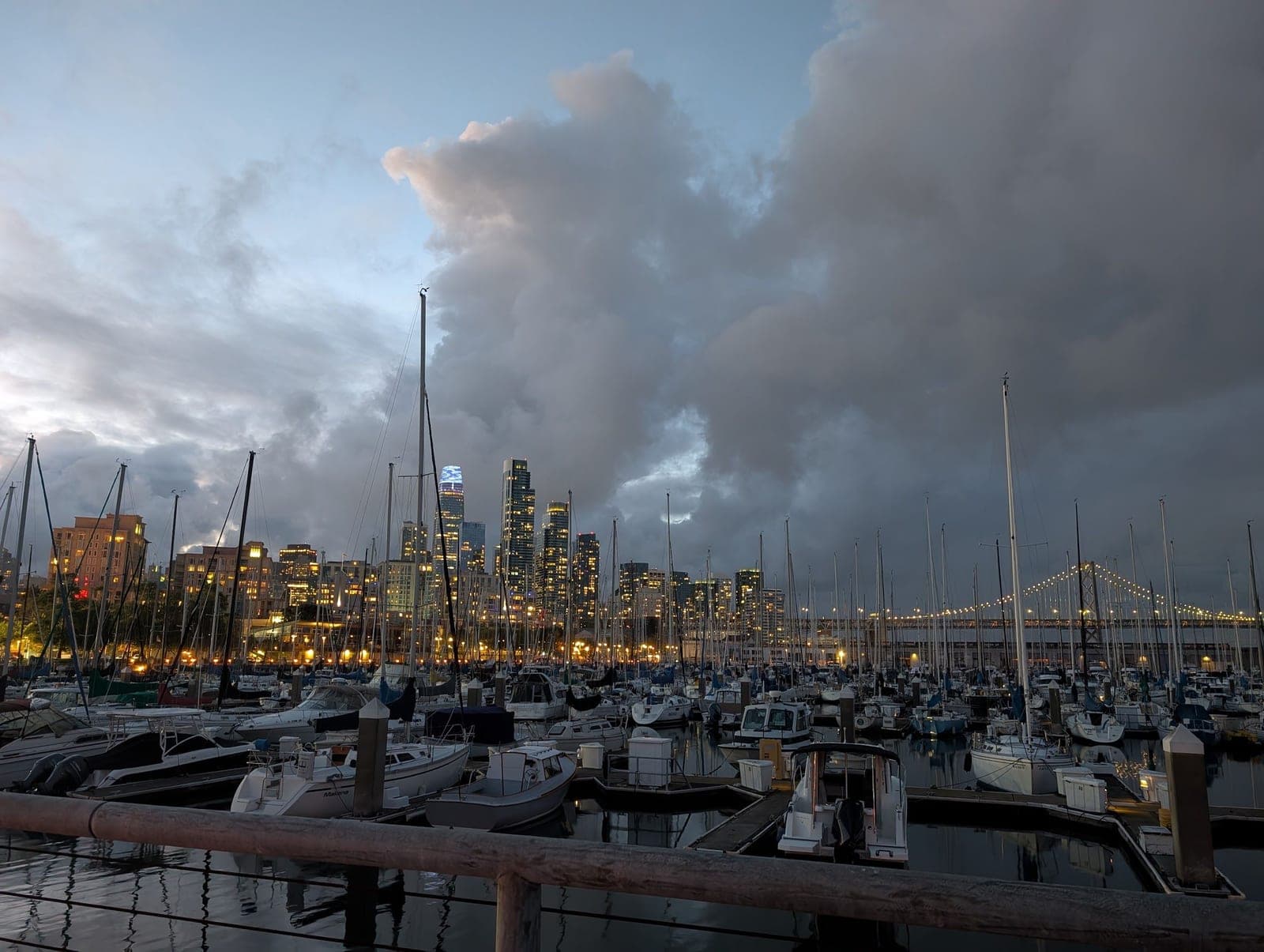 Sailboats in a marina at dusk, city lights and dark clouds behind.