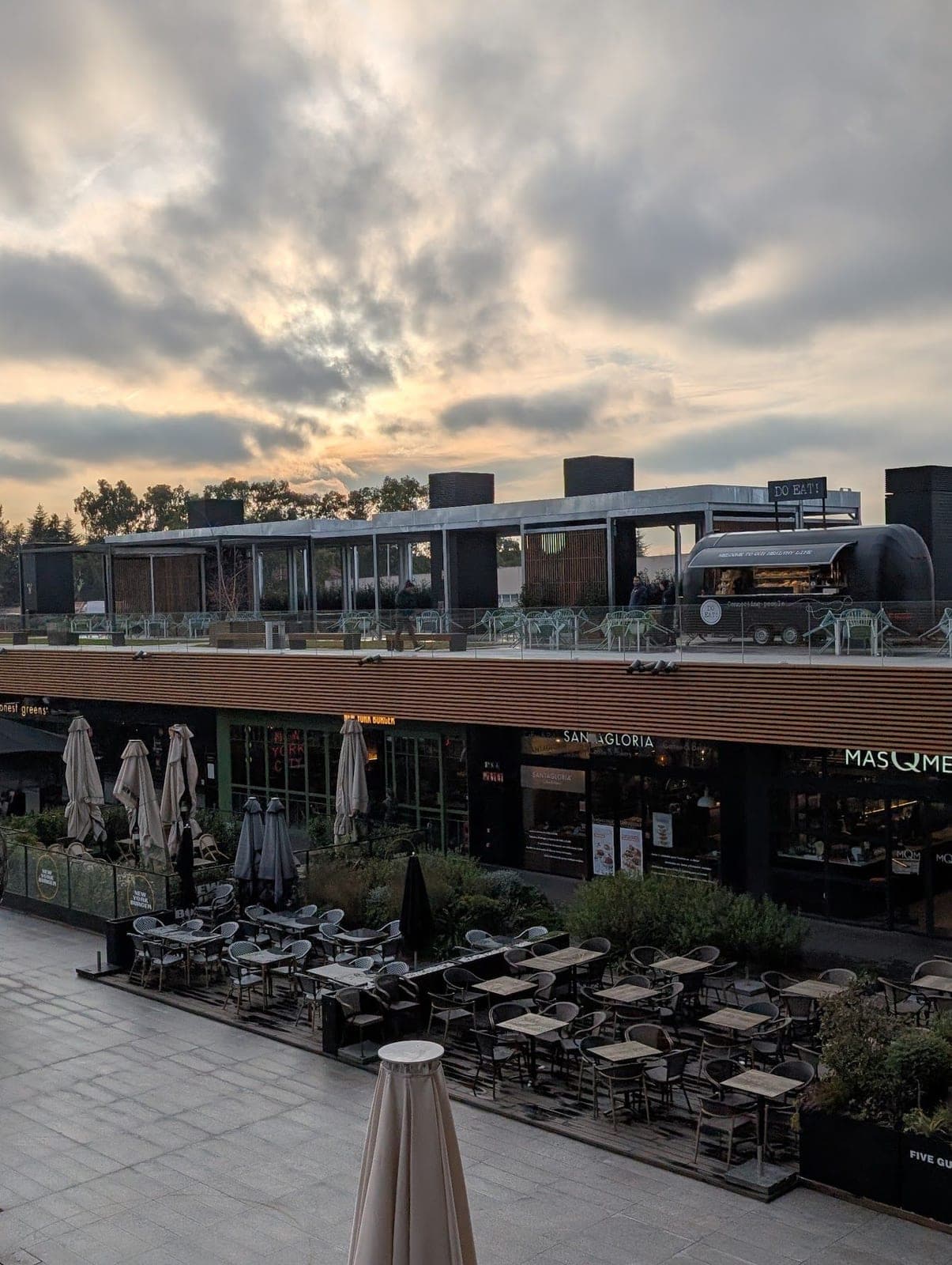 An empty restaurant terrace with folded umbrellas under a golden, cloudy sky.
