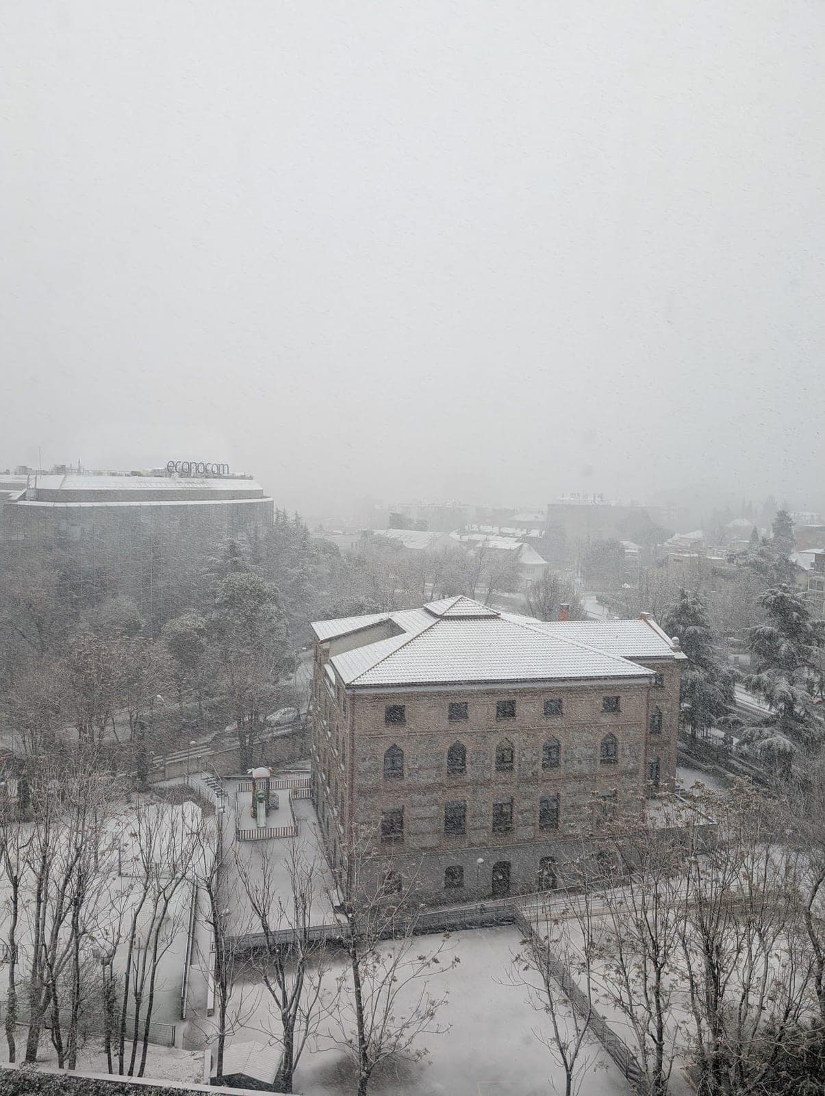 Snow over rooftops and a red brick building on a foggy morning.
