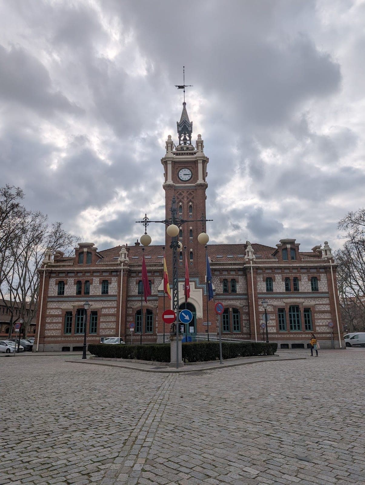 A red brick building with a tall clock tower and flags on a cobbled square under grey clouds.