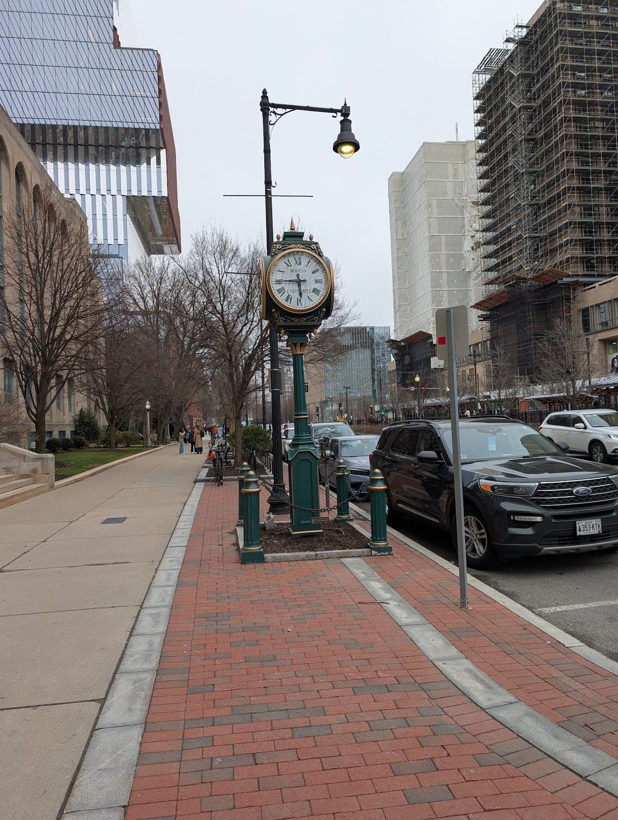 A brick sidewalk with a green street clock and lamp post, new buildings going up behind.