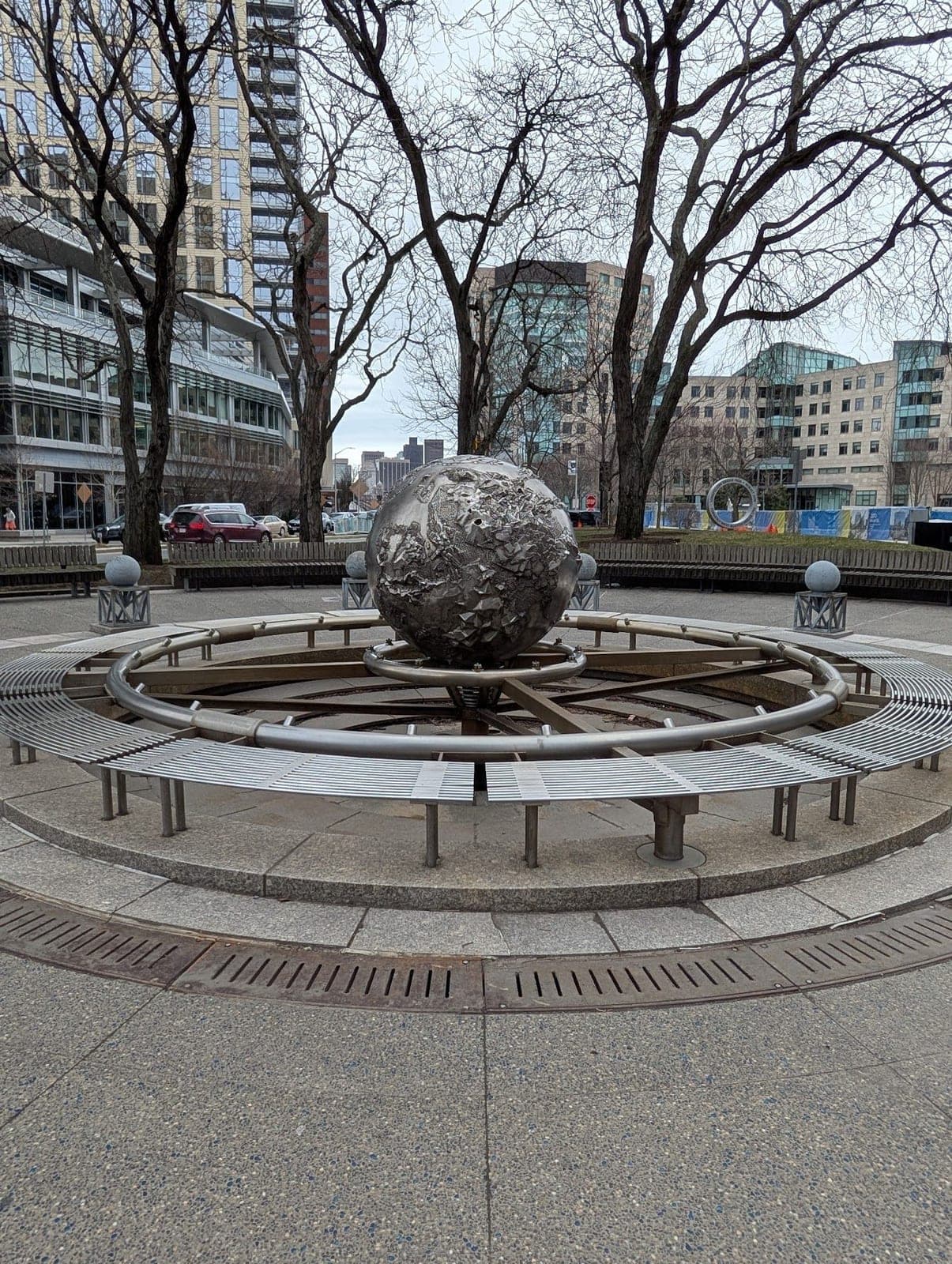 A metal globe sculpture at the centre of a round plaza of benches, bare trees behind.