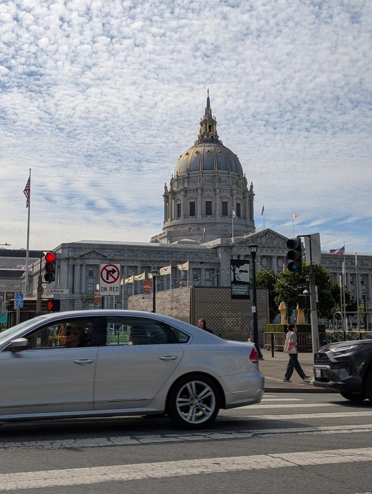 San Francisco City Hall's grey and gold dome under a sky full of little clouds, a car driving past.