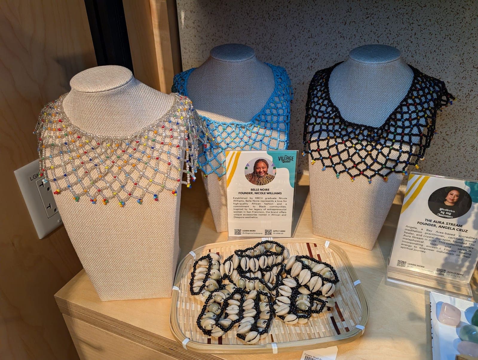 Beaded collars in white, blue and black on display busts, with cowrie shell bracelets in a basket.