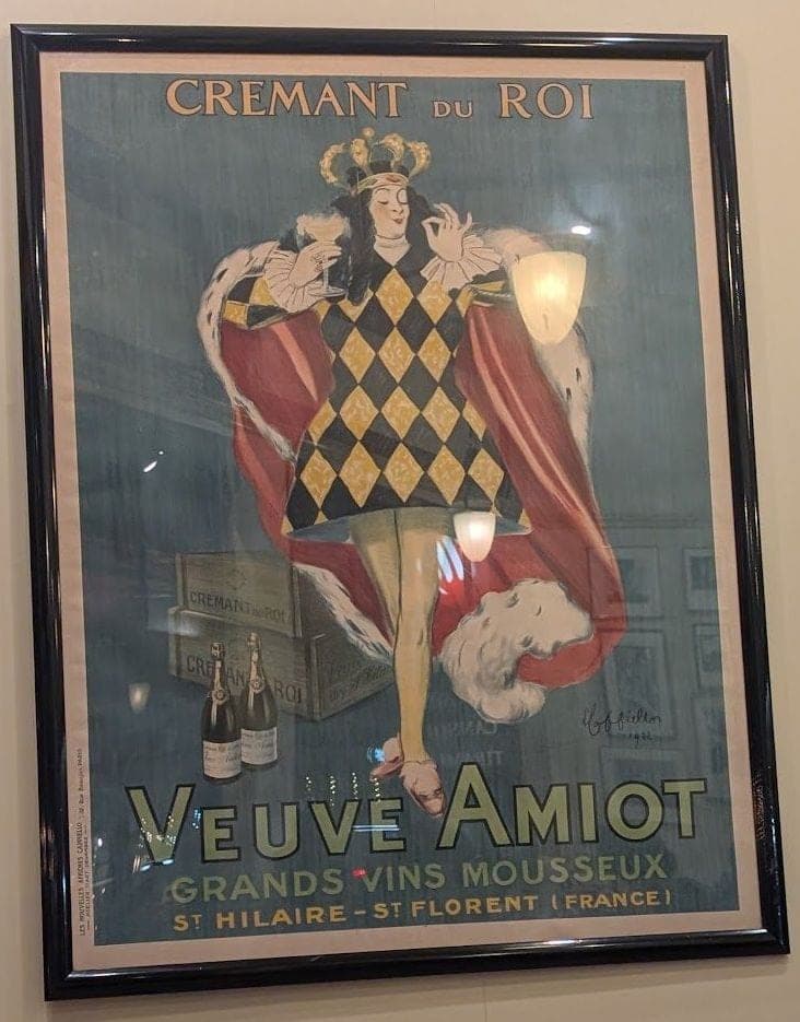 A framed 1922 champagne poster of a king in a harlequin costume and ermine cape, raising a glass: Veuve Amiot, Crémant du Roi.