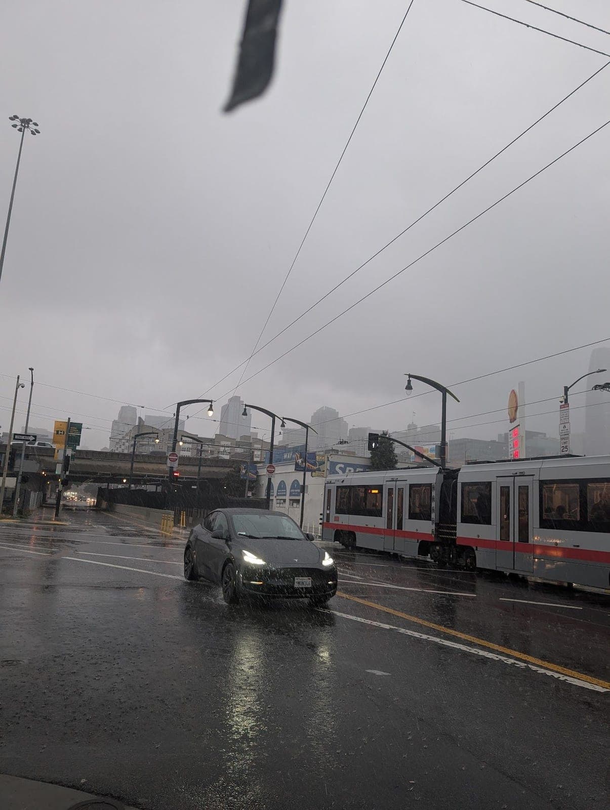 A rainy street with a car and a light-rail train under grey skies.