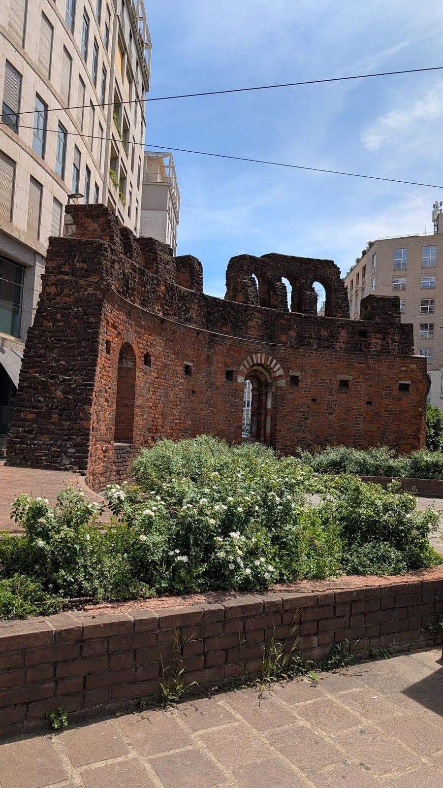 Ruined red brick walls with arches, surrounded by white flowers and modern apartment blocks.