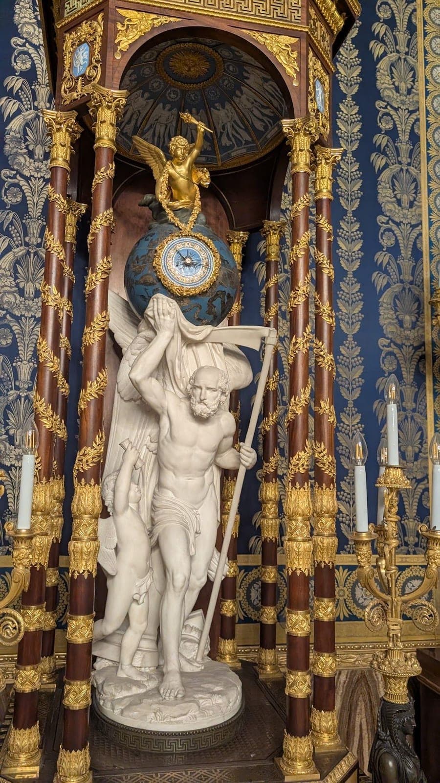 A white marble statue of Father Time with his scythe, holding up a blue and gold globe clock.