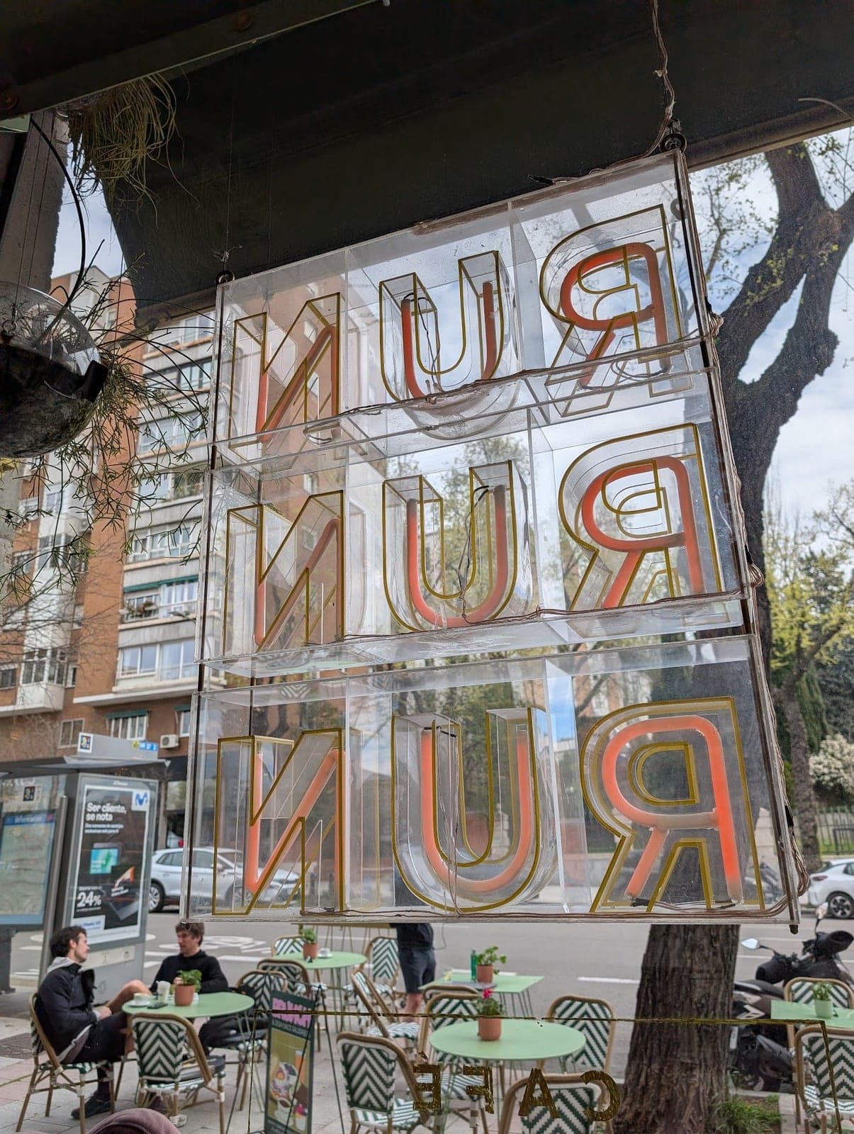 A neon sign spelling RUN three times, seen backwards from inside a café, green tables on the terrace outside.